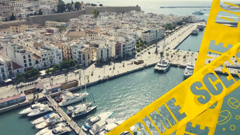 A scenic view of a marina in Ibiza with yachts docked along the waterfront, contrasted by yellow crime scene tape, suggesting a focus on safety or crime in the area.