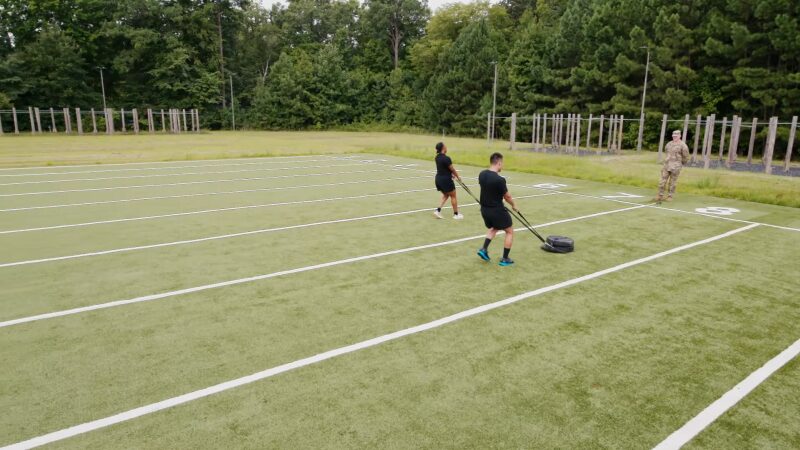 Two Soldiers Pull Weighted Sleds Across a Turf Field While a Military Instructor Observes, Demonstrating Strength Endurance and Work Capacity Training