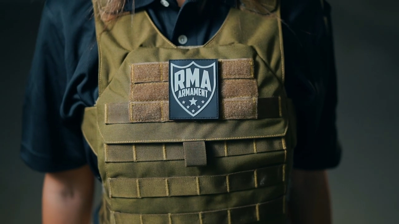 Close-up of a green bulletproof vest worn by a person, prominently displaying the RMA Armament logo on the front. The vest features multiple straps and compartments for added functionality