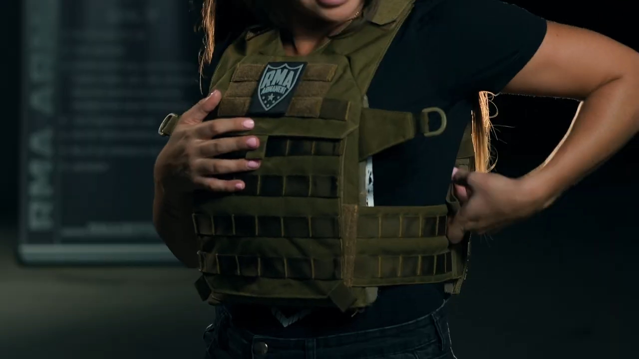 Close-up of a woman wearing a green bulletproof vest with a badge, adjusting the straps while standing in a dimly lit environment