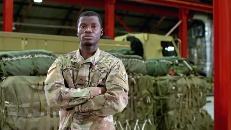 U.S. Army soldier in uniform standing confidently in a military warehouse with supplies in the background