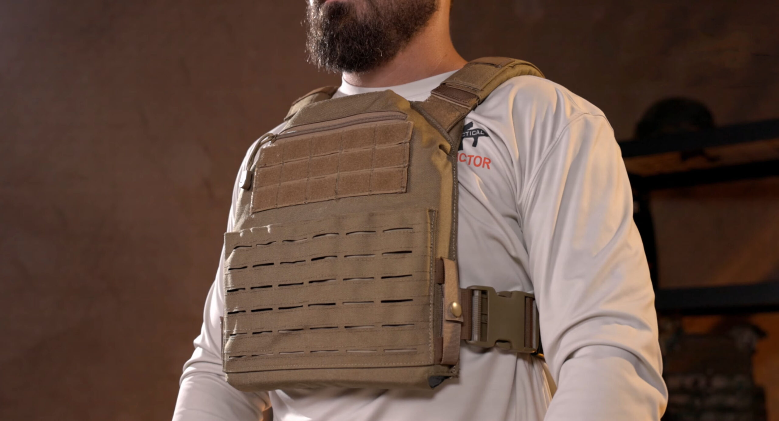 Close-up of a man wearing a tan tactical plate carrier over a white shirt in a rugged indoor setting