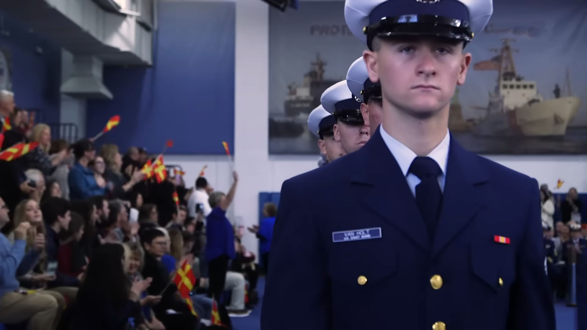 Coast Guard cadets in formal dress uniforms marching during a ceremony with a cheering crowd in the background