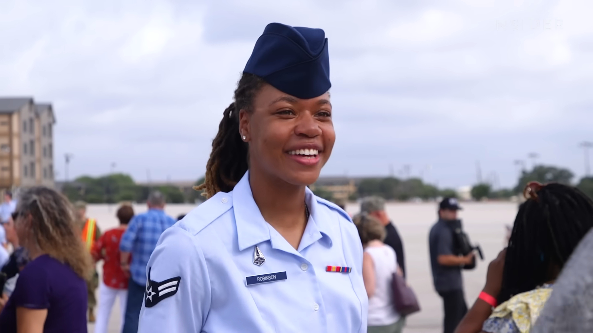 A U.S. Air Force member in uniform, smiling outdoors during a graduation event with people and buildings in the background