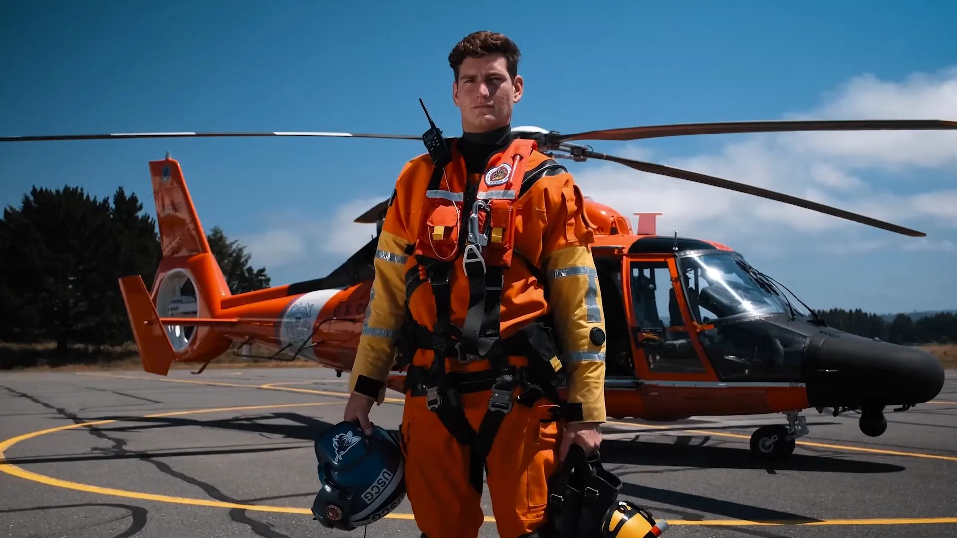 A U.S. Coast Guard rescue swimmer in an orange suit stands in front of a Coast Guard helicopter holding gear