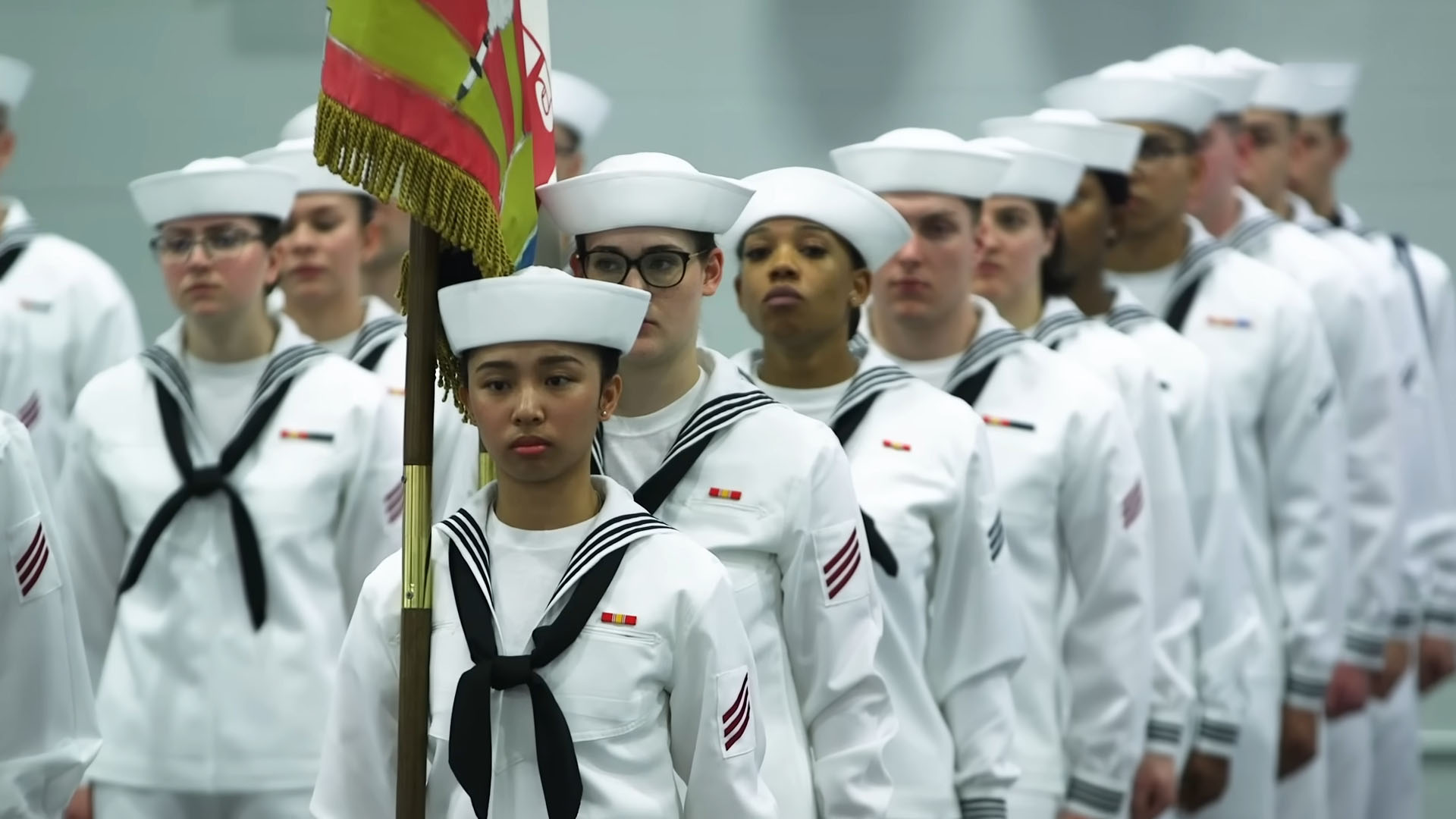 Group of U.S. Navy recruits dressed in white uniforms standing in formation, with a recruit holding a flag at the front