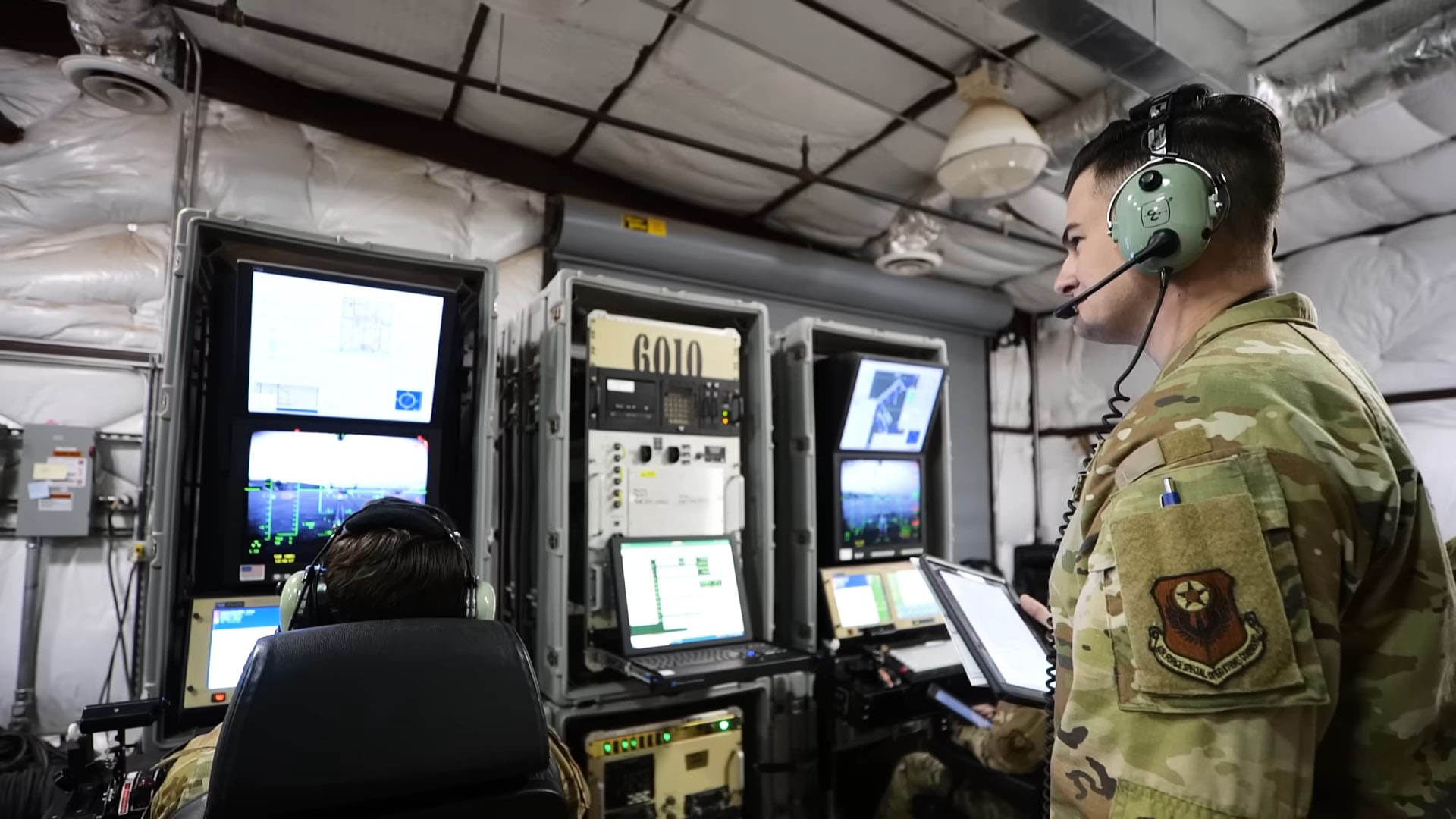 Military personnel in a control room with multiple screens and equipment, overseeing UAV operations, wearing headsets for communication