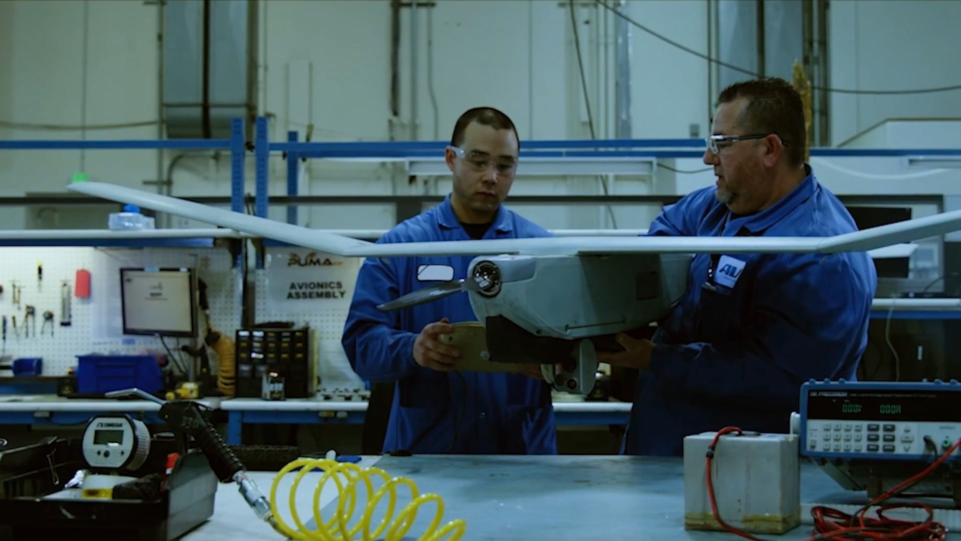 Two engineers in blue lab coats working on a drone in a workshop filled with tools and equipment