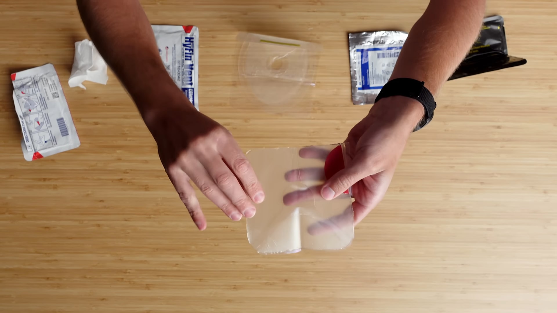 Hands holding a chest seal with medical supplies placed on a wooden table in the background