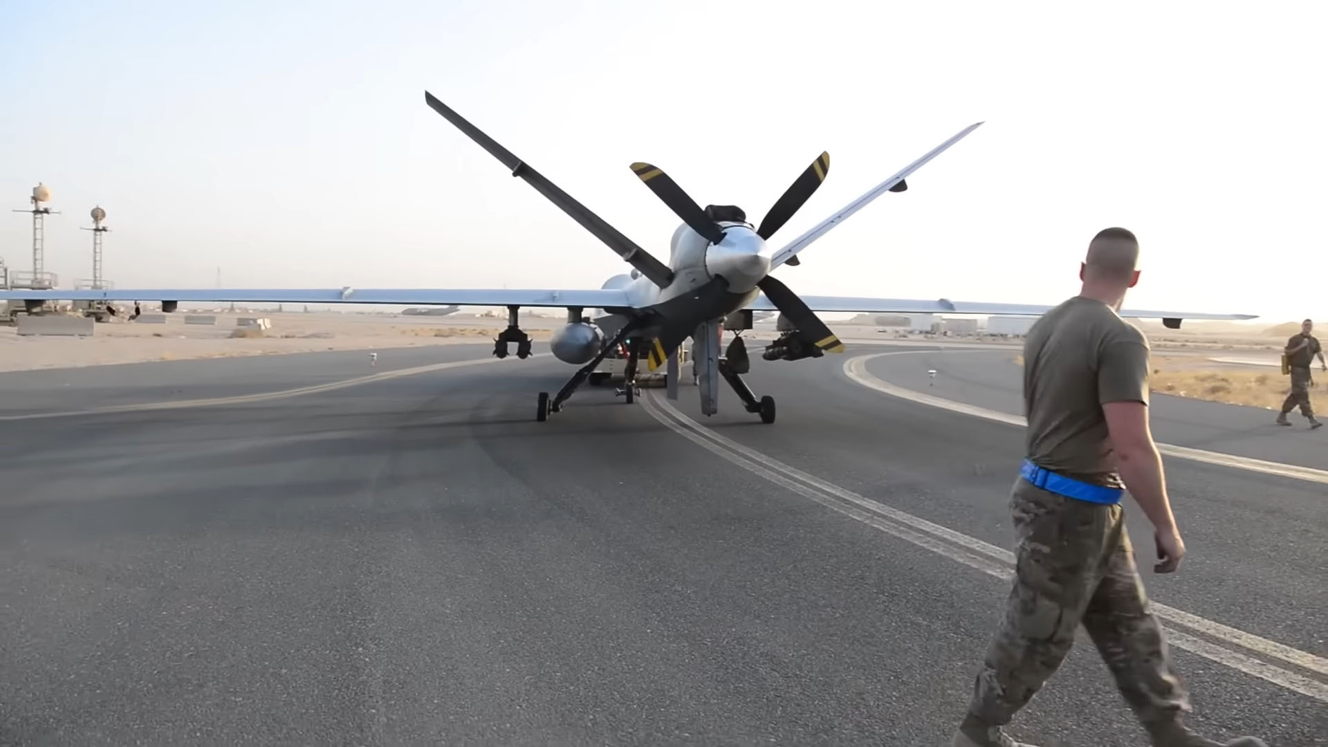 Military personnel walking near a large drone on an airstrip in a desert environment