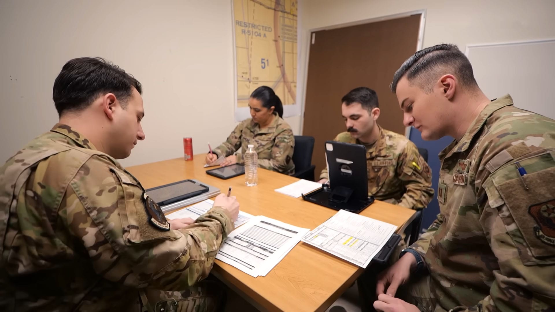 A group of military personnel in camouflage uniforms sitting around a table, reviewing documents and electronic devices during a mission briefing