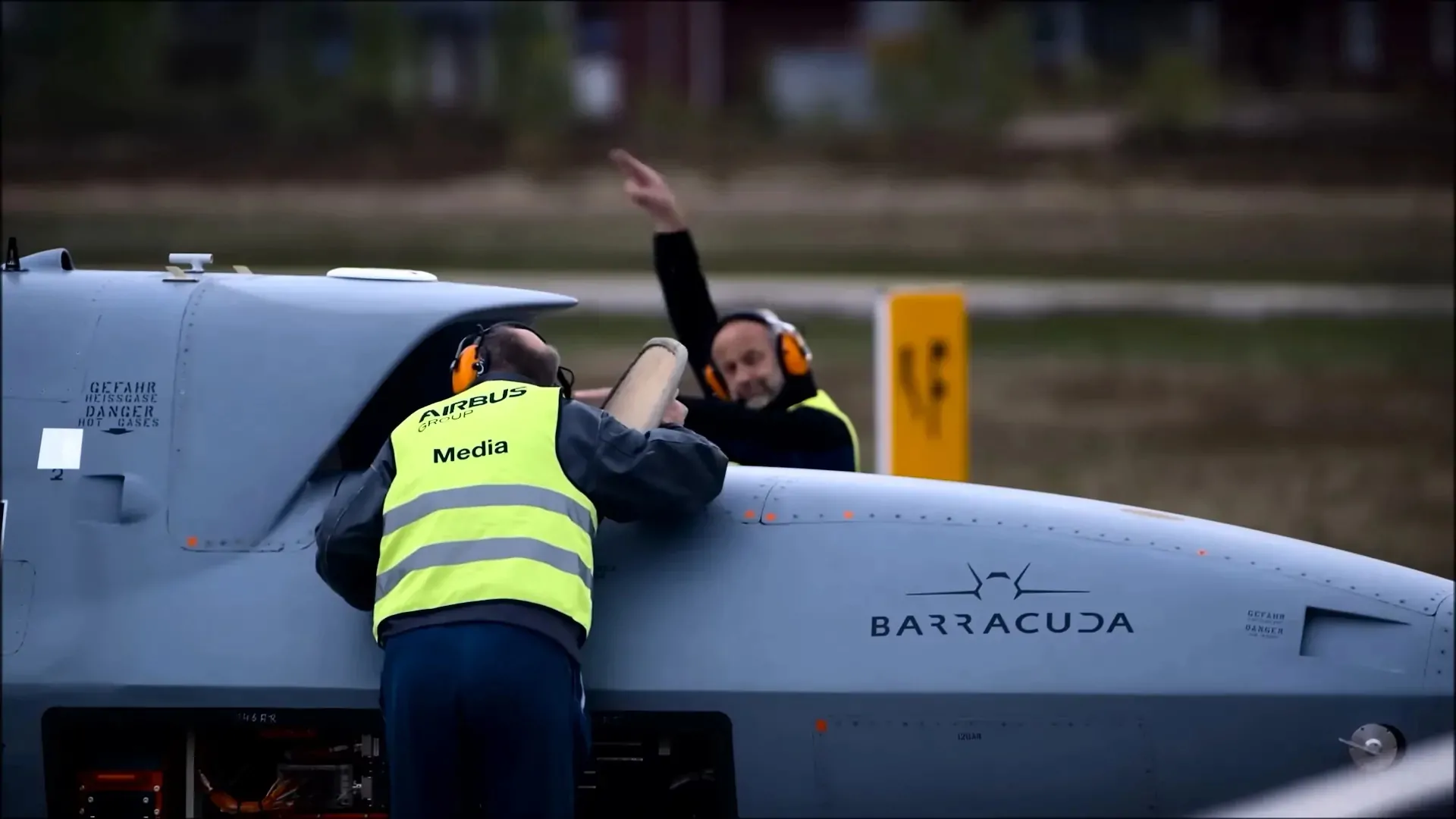 Two ground crew members in high-visibility vests performing pre-flight checks on the Barracuda UAV, with one signaling