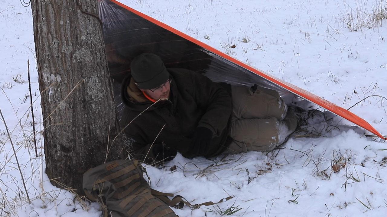 A person lying under an emergency blanket shelter tied to a tree in a snowy field, with a backpack nearby