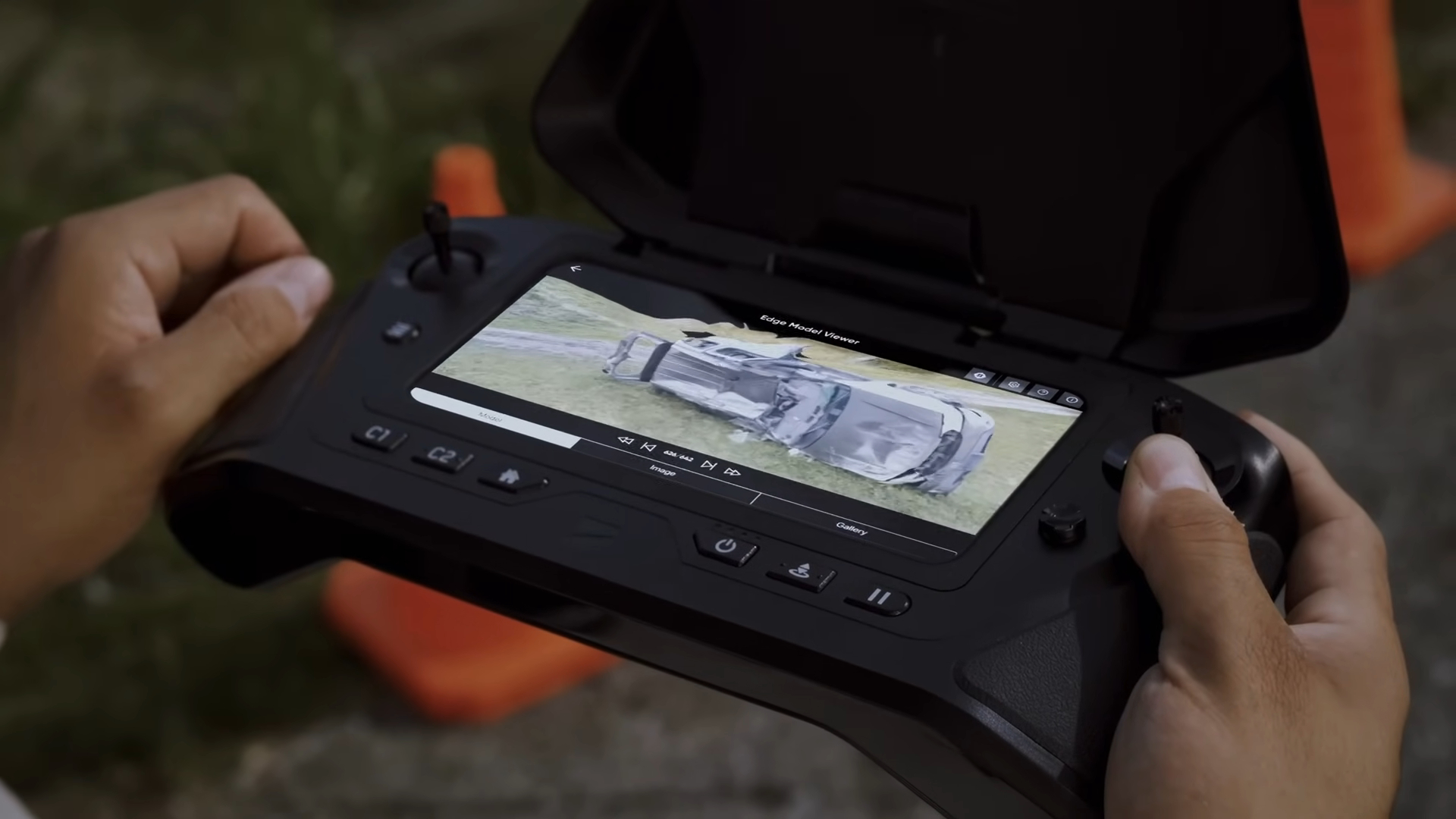 Hands holding a drone controller displaying a high-resolution image of a flipped vehicle on the screen