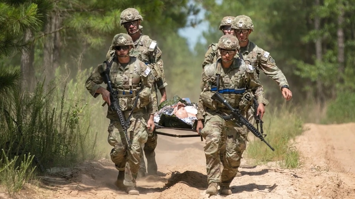 Soldiers in full gear carrying a wounded comrade on a stretcher through a forest training ground