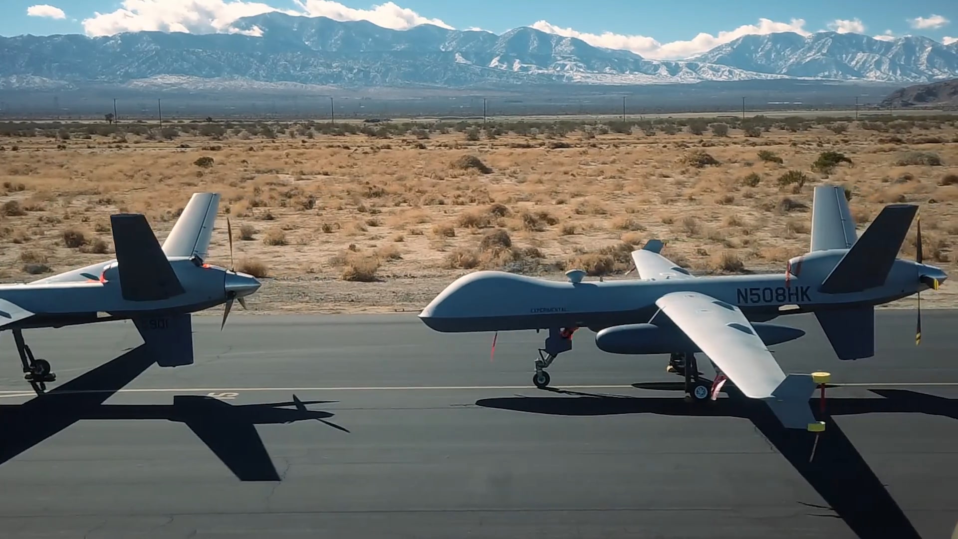 Two military drones parked on a runway with a desert backdrop and snow-capped mountains in the distance