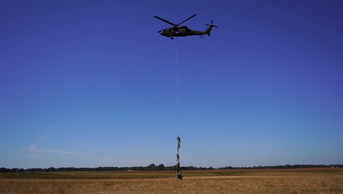 A military helicopter hovers in the clear blue sky while a group of soldiers dangles from a long rope, performing a fast-rope insertion onto an open field