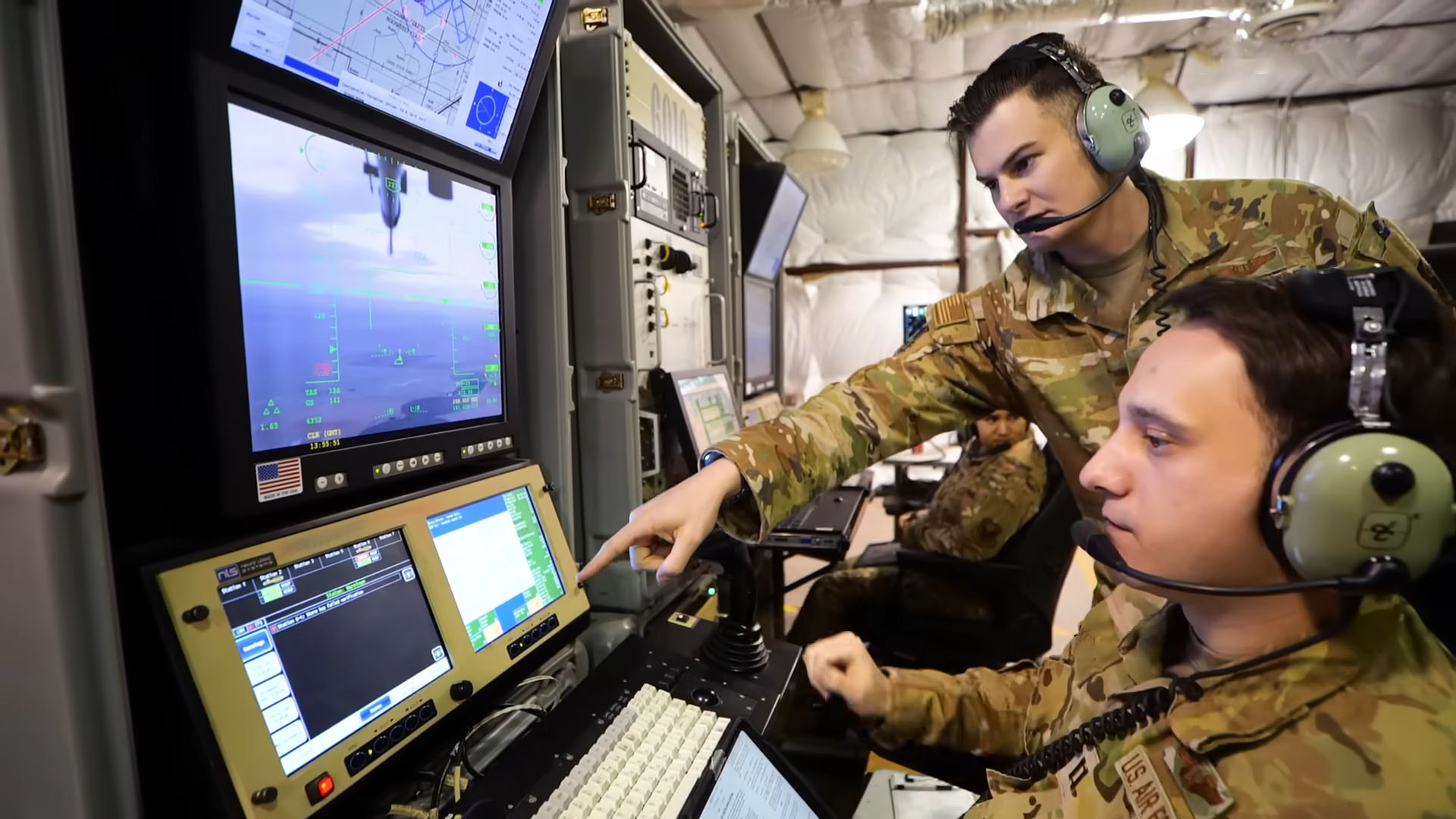 Two military personnel in camouflage uniforms operating a UAV control station with multiple screens and controls, wearing headsets