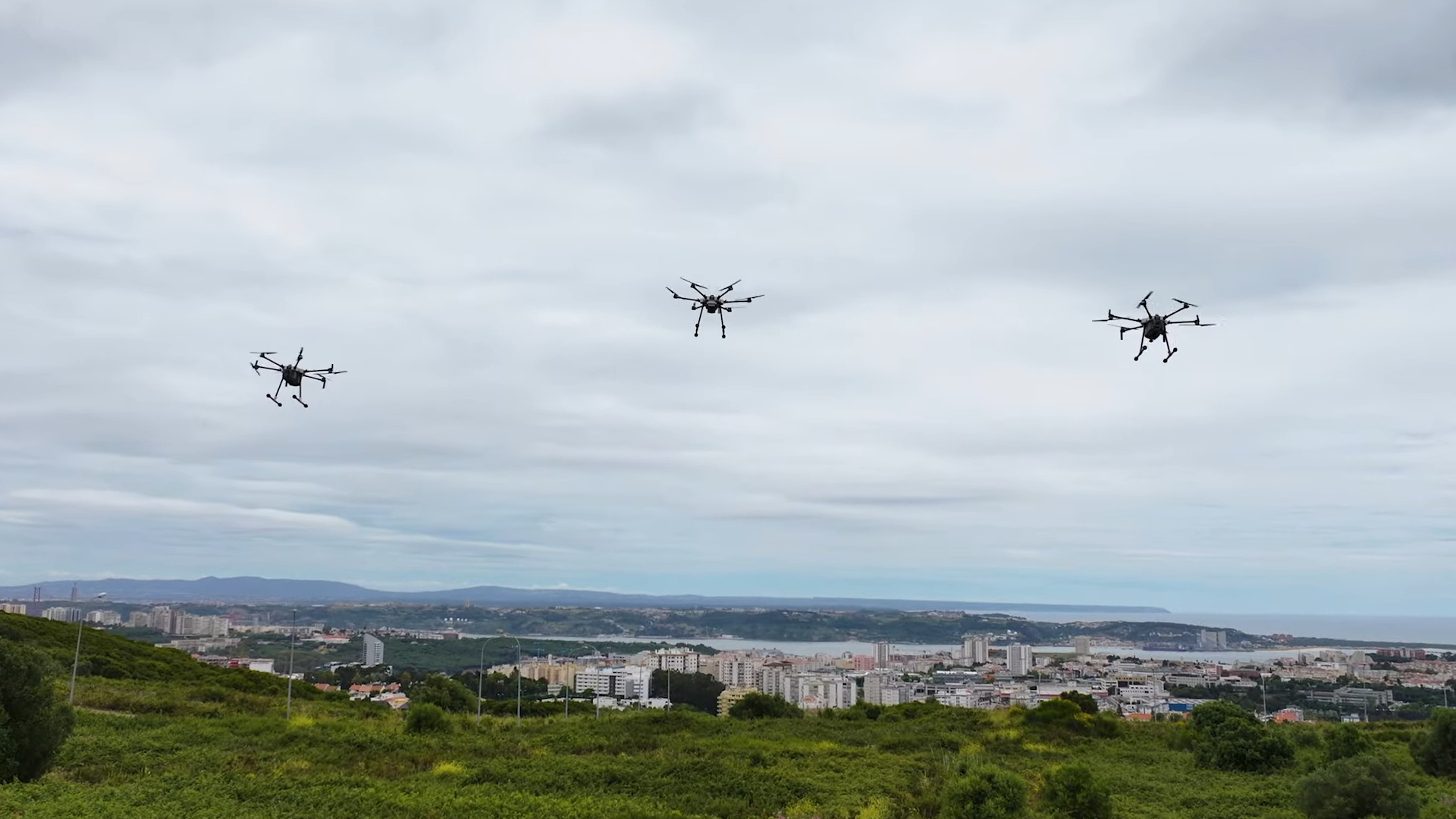 Three drones flying in formation over a green landscape with a city and ocean visible in the background under cloudy skies