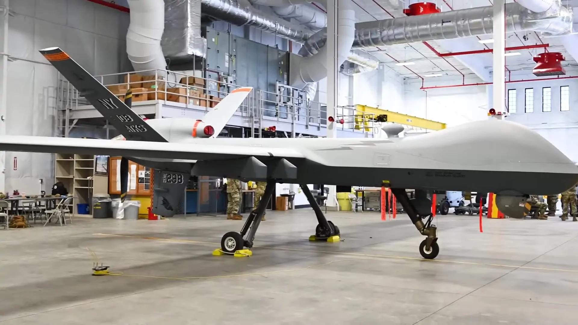 A large MQ-9 Reaper drone inside an industrial hangar, with military personnel in the background