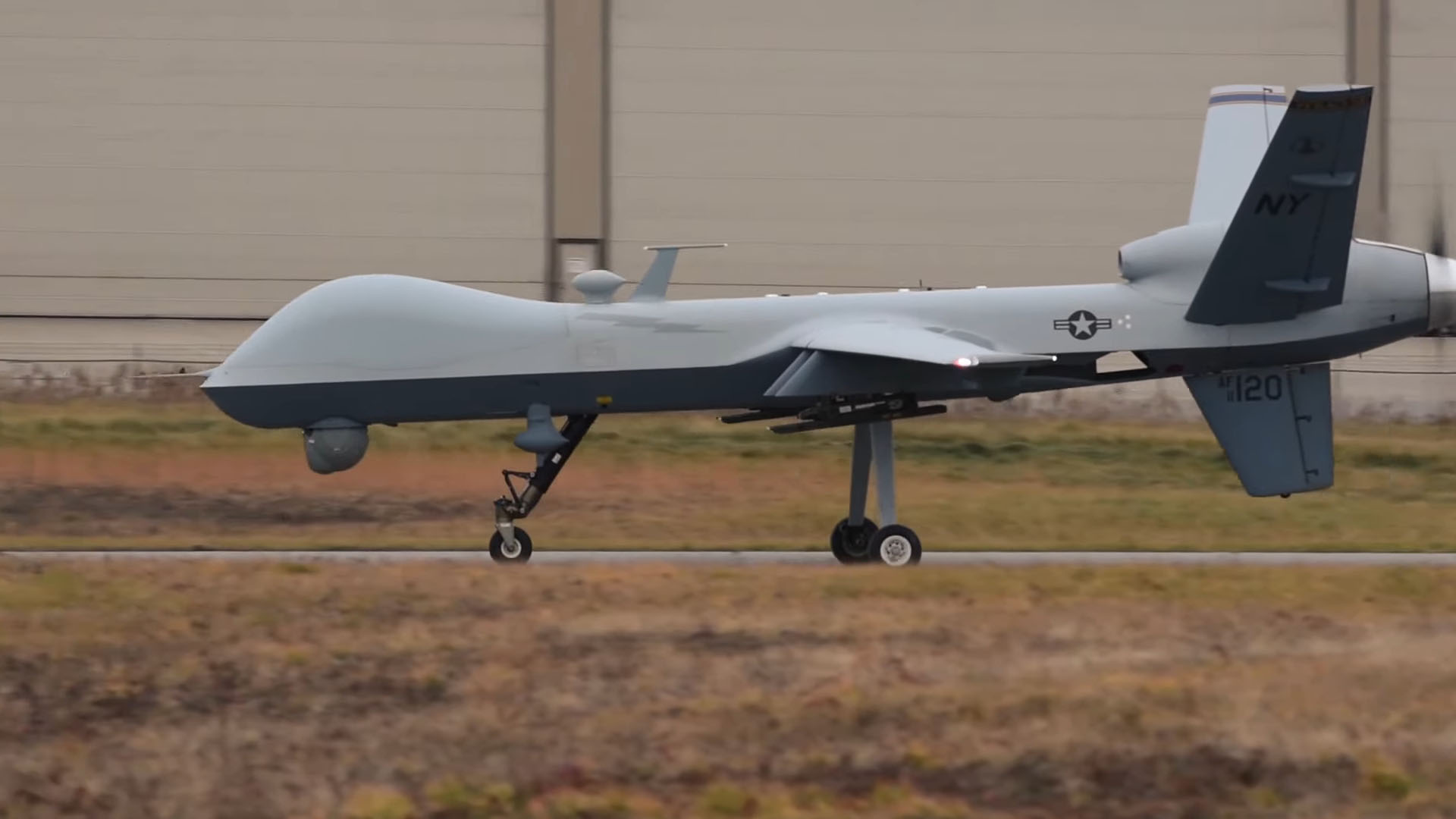 MQ-9 Reaper drone preparing for takeoff on a runway, with a beige building in the background