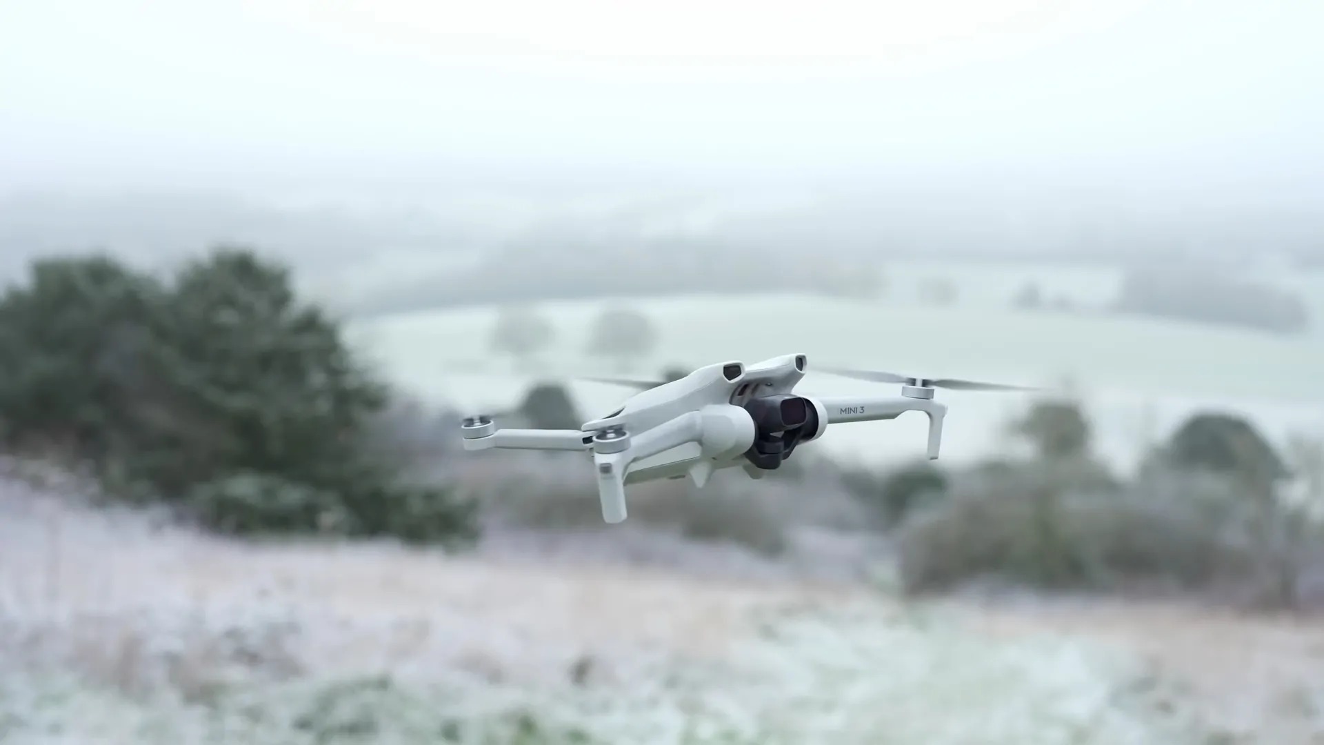 A white DJI Mini 3 drone hovering over a frosty landscape with blurred trees and fields in the background