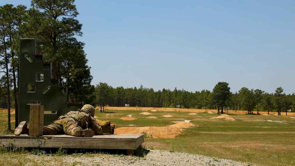 A soldier in camouflage lying on a wooden platform, aiming at a distant target in an outdoor shooting range