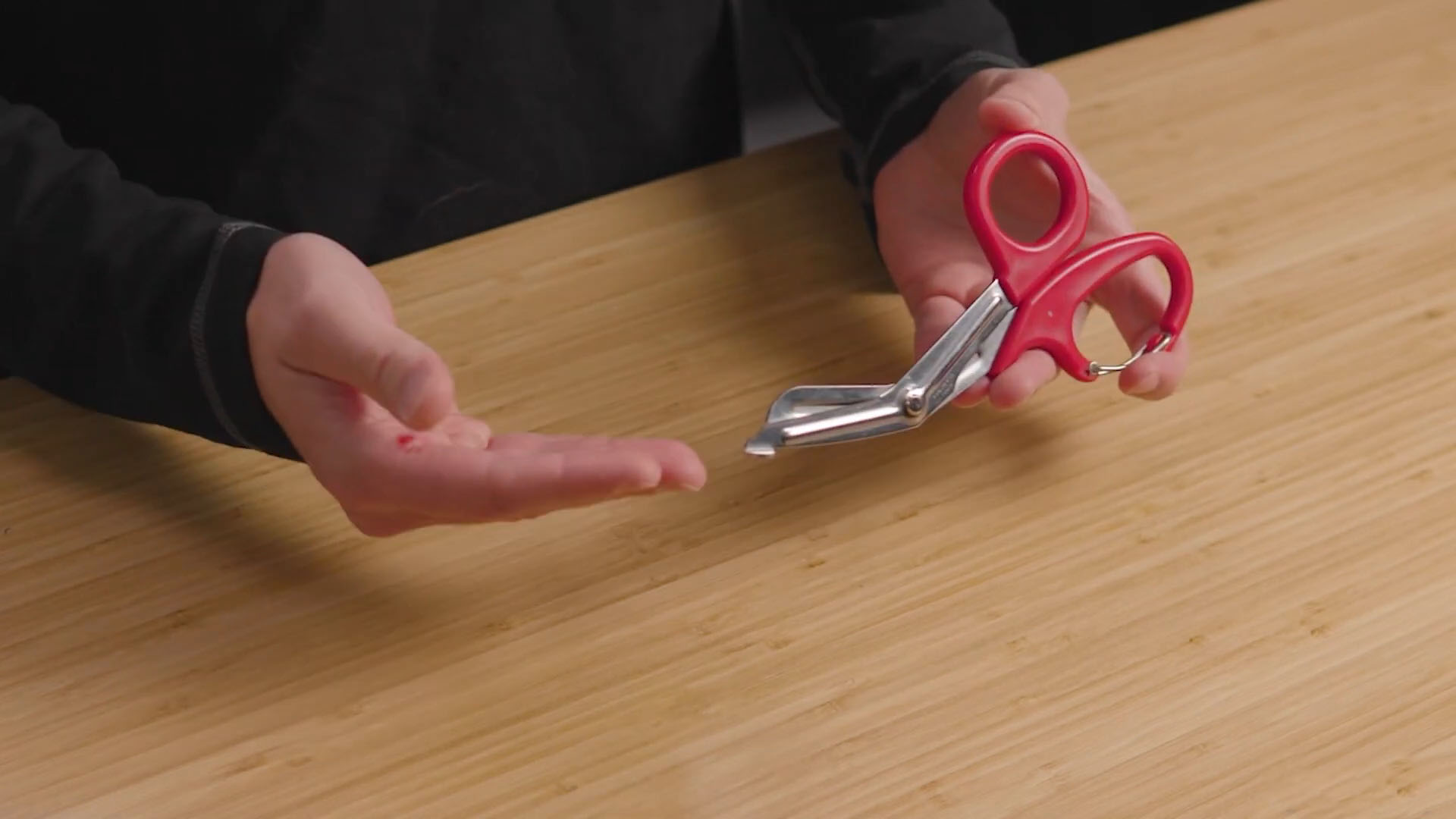 Hands holding red-handled trauma shears on a wooden surface, demonstrating their design