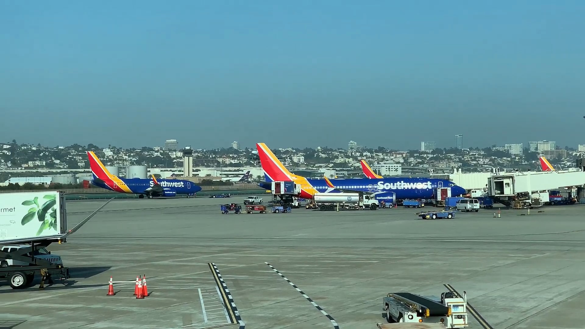 Multiple Southwest Airlines planes parked at the gates of an airport with ground crew and fueling trucks servicing them. A city skyline is visible in the background.