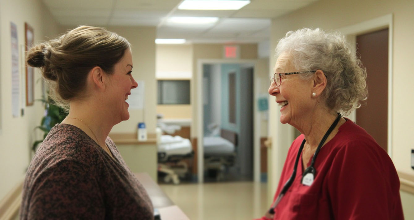 A nurse in red scrubs smiling and talking with a woman in a medical facility hallway, with hospital beds visible in the background