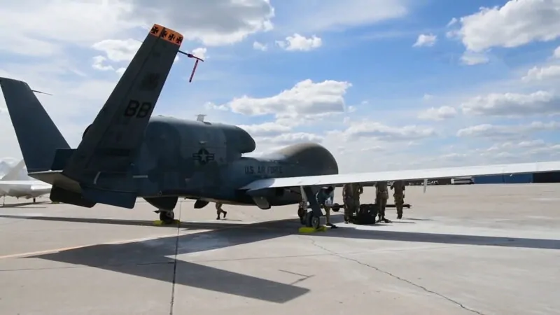 An RQ-4 Global Hawk drone stationed on a runway with U.S. Air Force personnel preparing for operations under a partly cloudy sky