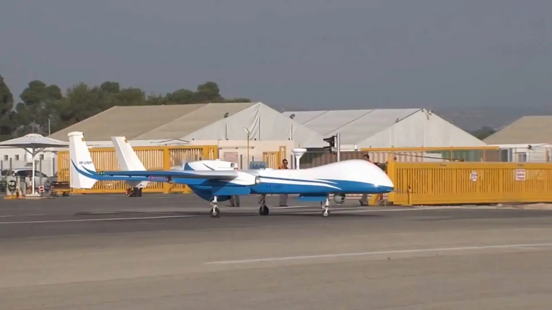 A Heron TP unmanned aerial vehicle (UAV) painted in blue and white, parked on an airfield with tents and security barriers in the background