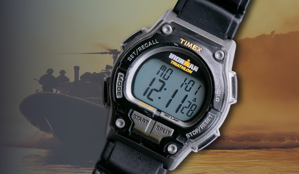 A rugged Timex Ironman Triathlon digital watch displayed against a military boat and helicopter background